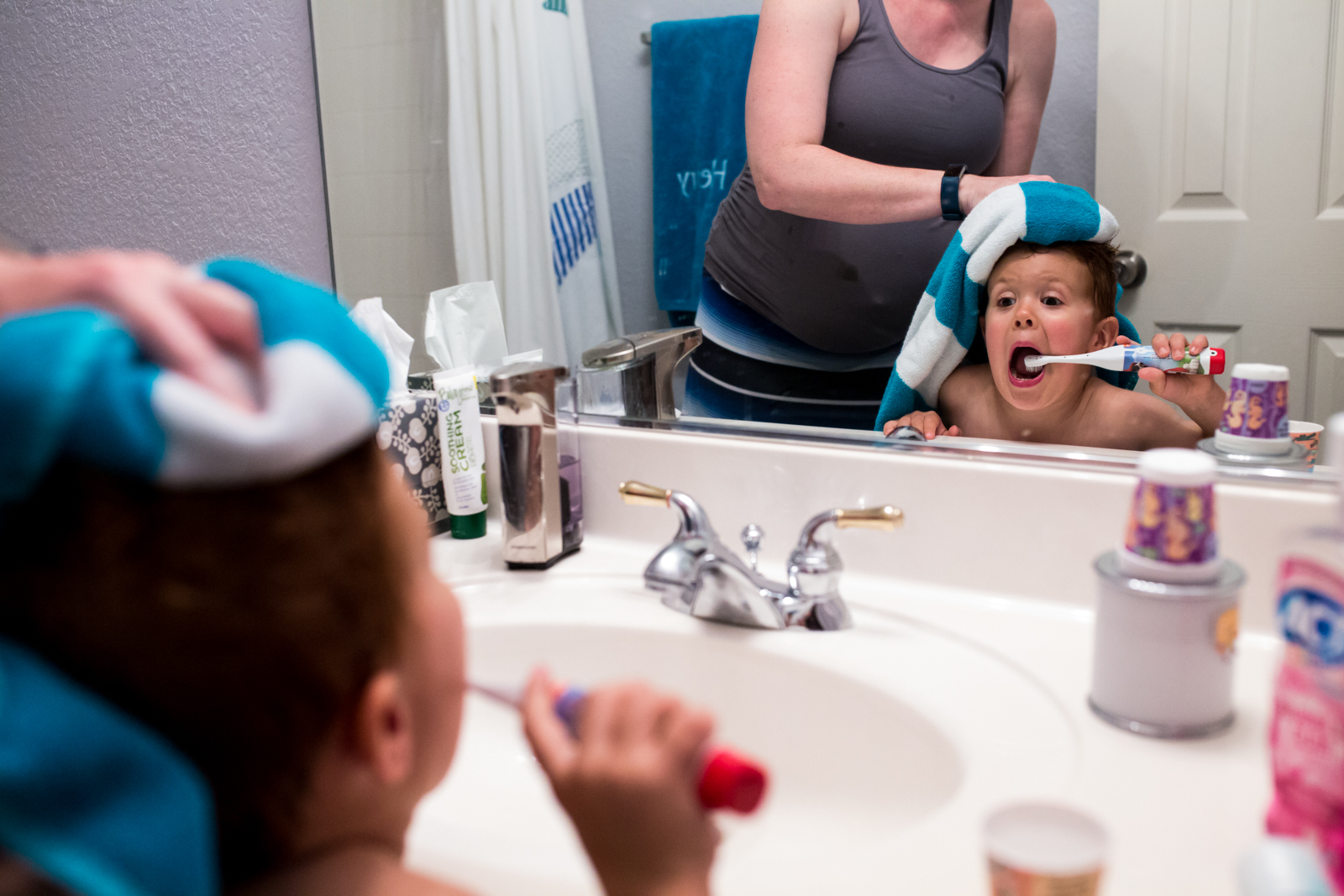 Little boy makes a goofy face in the mirror while his mom tries to dry off his hair after a bath, photo taken by Lawren Rose Photography in McKinney Texas