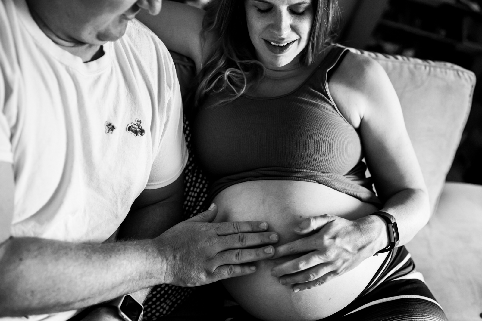 Lawren Rose Photography takes a photo of a husband and pregnant wife sitting on a couch admiring her pregnant belly