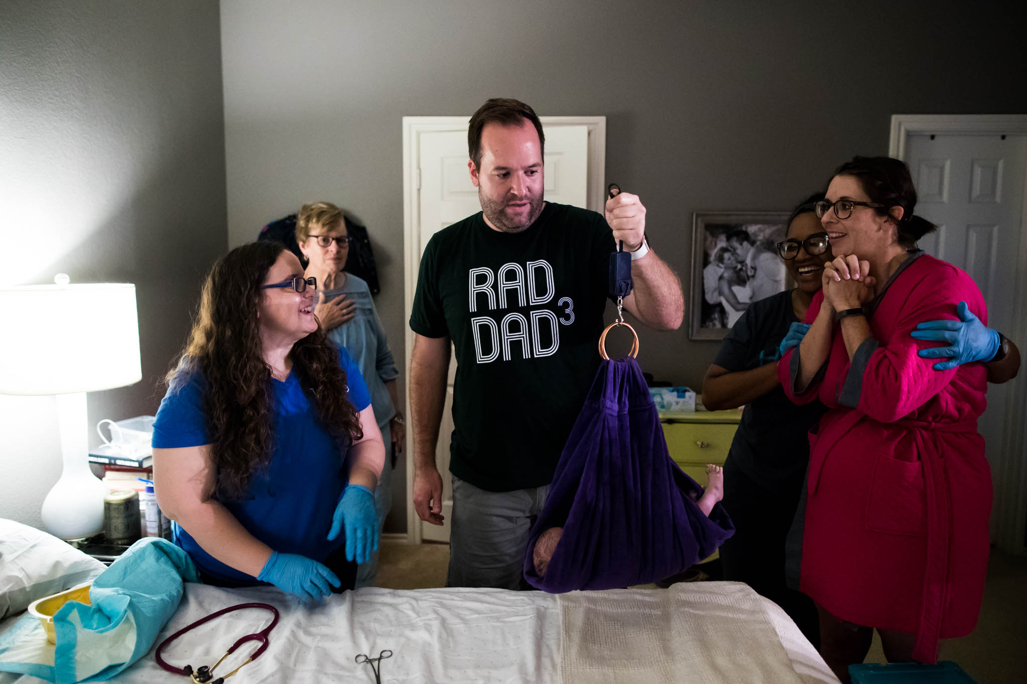 Dallas Birth Photographer Lawren Rose Photography takes an image of the birth team and family surrounding the new Dad holding up the handheld scale to weigh his new baby right after their successful home birth